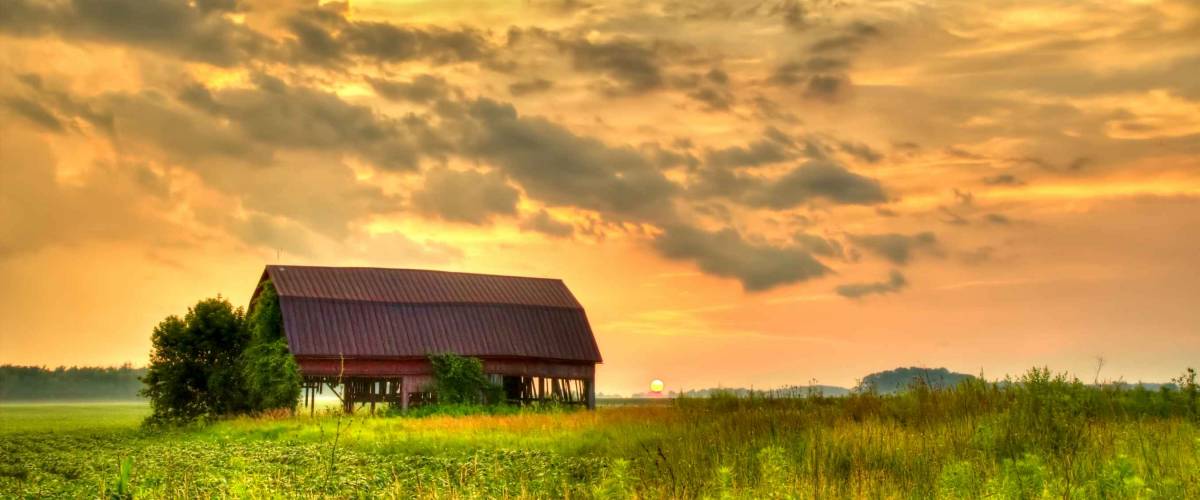 American Midwest Barn Landscape. Sunset over a farm field with a traditional red barn at the horizon.